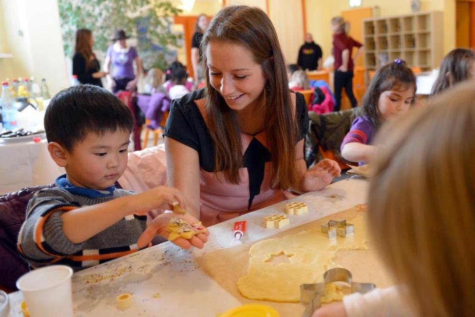 A young female volunteer sits at a table with some children and assists with baking cookies 